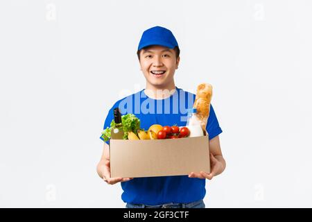 Online-Shopping, Lebensmittelzustellung und Versandkonzept. Freundlich lächelnder junger asiatischer Kurier in blauer Uniform mit Lebensmitteln, bringen Sie Ordnung zu Stockfoto