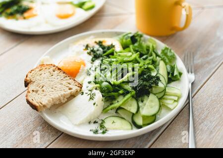 Spiegeleier mit grünem Salat aus Gurke und Pfeffer auf Holztisch Hintergrund, leckeres Frühstück Stockfoto