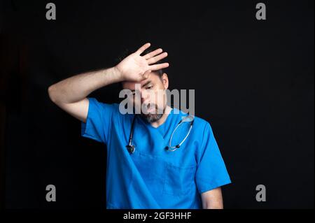 Arzt in blauer Krankenhausuniform, dunkel und bärtig, trägt ein Stethoskop um seinen Hals und wischt sich mit der Hand mit einem müden OP die Stirn Stockfoto