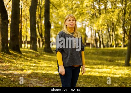 Defokussieren Sie eine ernsthafte blonde Frau aus den 40er Jahren, die im gelben Herbstpark steht. Frohe, schöne Dame. Frauen tragen grauen Pullover, gelben Rollkragen, Anhänger, Jeans Stockfoto