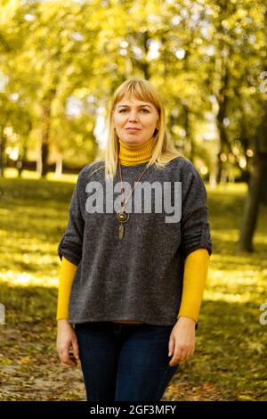 Defokussieren Licht lächelnde blonde 40er Frau im gelben Herbst Park stehen. Frohe, schöne Dame. Frauen tragen grauen Pullover, gelben Rollkragen, Anhänger Stockfoto