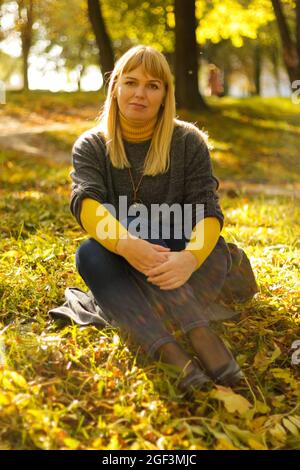 Defokussieren Sie lächelnde blonde Frau aus den 40er Jahren, die im gelben Herbstpark sitzt. Frohe, schöne Dame. Frauen tragen grauen Pullover, gelben Rollkragen, Anhänger, Jeans Stockfoto
