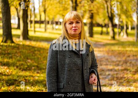 Unschärfe-Außenportrait der positiven selbstbewussten reifen Frau. Lächelnde weibliche Blondine. Elegante Frau mittleren Alters im Herbstpark. Nicht fokussiert Stockfoto