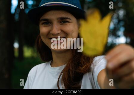 Unschärfe-Porträt einer glücklich lächelnden Frau mit braunen Haaren trägt einen Hut im Freien. Weibliche Hand mit gelbem, trockenem Blatt. Hallo Herbst. Frauen stehen an Stockfoto
