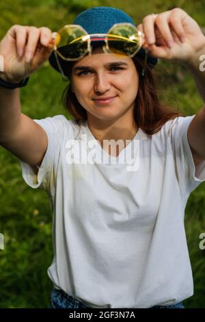 Unschärfe-Nahaufnahme Porträt einer glücklichen jungen Frau mit braunen Haaren trägt einen Hut im Freien. Gelbe Sonnenbrille. Grüne Natur Hintergrund. Suchen Stockfoto