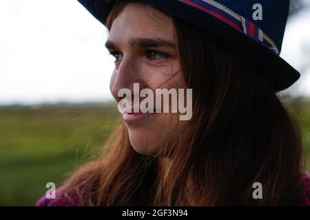 Unschärfe-Nahaufnahme Porträt einer glücklichen jungen Frau mit braunen Haaren trägt einen Hut im Freien. Grüne Natur Hintergrund. Wegschauen. Nicht fokussiert Stockfoto