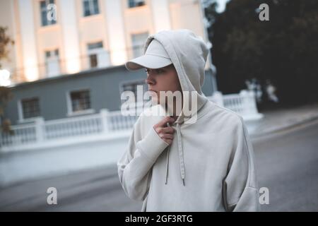 Junge Teenager, 18-19 Jahre alt, tragen trendigen beigefarbenen Hoodie und Mütze, trainieren früh morgens im Dunkeln über dem Stadthintergrund. Selektiver Fokus. Teenagerhoo Stockfoto