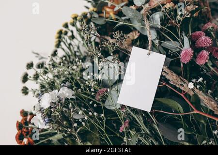 Moody feminine Hochzeit, Geburtstag Mockup Szene. Nahaufnahme einer leeren Papierkarte, Geschenketikett auf einem bunten Blumenstrauß. Gypsophila und bunte santini Stockfoto