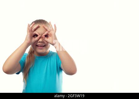 Fröhliches kaukasisches kleines Mädchen in einem blauen T-Shirt macht mit ihren Fingern eine Brille auf weißem Hintergrund. Stockfoto