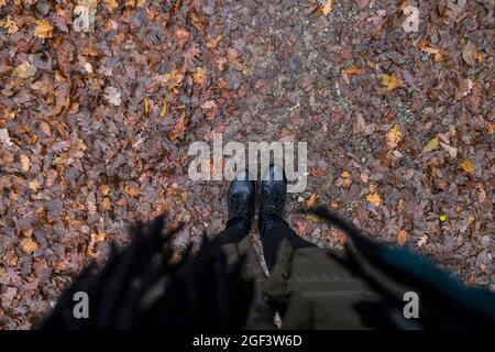 Weibliche schwarze Lederstiefel, Mantel und Schal einer jungen Frau, die auf dem Boden steht und mit herbstlichen Herbstblättern bedeckt ist. Herbststimmung Stockfoto