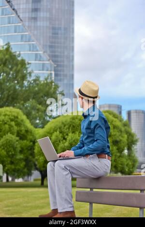 Ein Mann mit einem Laptop arbeitet ferngesteuert auf einer Bank im Freien und schaut auf ein Bürogebäude. Stockfoto