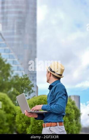 Ein junger Mann mit einem Laptop arbeitet fernab von draußen und blickt auf das Bürogebäude. Aufgrund von Einschränkungen während einer Pandemie Remote arbeiten und studieren. Stockfoto
