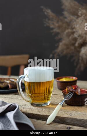 Glas mit Bier und Schaum und Gabel mit einem Stück Wurst und Ketchup auf Holzboden, gebratene Würstchen auf einem Teller und Ketchup mit Senf Stockfoto