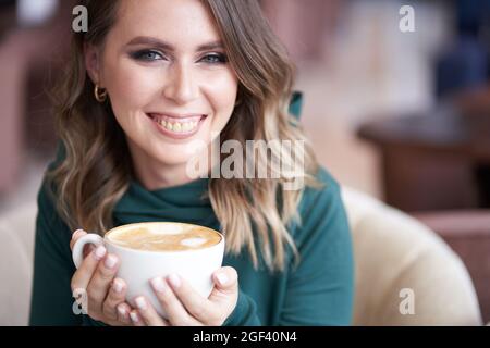 Eine glückliche, schöne Brünette mit einer schönen Maniküre, die eine Tasse Kaffee in den Händen hält, einen grünen Rollkragen trägt, in einem Café sitzt und direkt in die Kamera schaut. Hochwertige Fotos Stockfoto