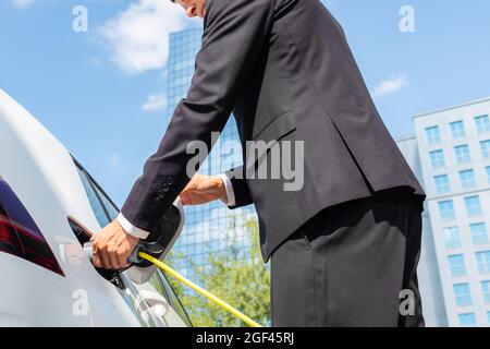 Mann in einem schwarzen Anzug, der in der Nähe eines Elektroautos steht, das lädt, und die Zeit auf einem Smartphone anpasst Stockfoto