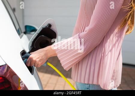 Nahaufnahme einer kaukasischen weiblichen Hand, die ein Elektroauto mit eingestecktem Netzkabel lädt Stockfoto