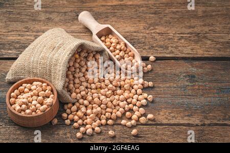 Trockene, rohe Kichererbsen in einem Leinenbeutel, einer Holzlöffel und einer Schüssel auf Holzboden. Seitenansicht, Platz zum Kopieren. Stockfoto