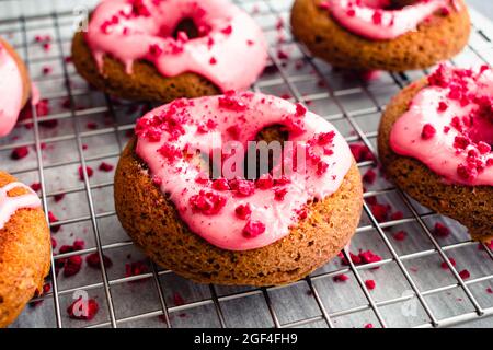 Himbeer-gebackene Donuts mit Himbeer-Glasur auf einem Wire Cooling Rack: Gebackene Donuts mit rosa Himbeer-Glasur und roten Streuseln Stockfoto