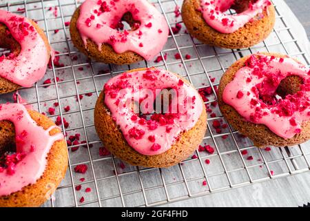 Himbeer-gebackene Donuts mit Himbeer-Glasur auf einem Wire Cooling Rack: Gebackene Donuts mit rosa Himbeer-Glasur und roten Streuseln Stockfoto