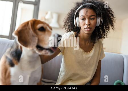 Junge afrikanerin mit Kopfhörern, die sich mit ihrem Hund zu Hause auf der Couch entspannt und kuschelt Stockfoto