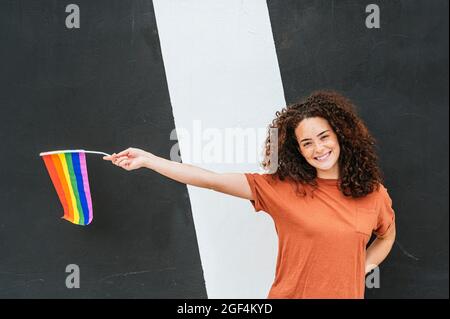 Lächelnde junge Frau, die mit einer Regenbogenfahne vor einer schwarz-weißen Wand steht Stockfoto