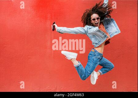 Aufgeregte junge Frau mit Sonnenbrille, die vor der roten Wand springt Stockfoto