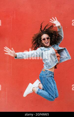 Fröhliche Frau mit Sonnenbrille, die vor der roten Wand springt Stockfoto