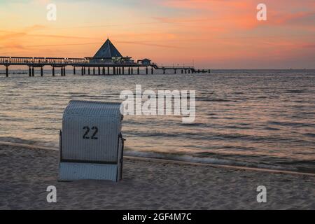 Deutschland, Mecklenburg-Vorpommern, Heringsdorf, Liegestuhl mit Kapuze, der in der Abenddämmerung am sandigen Küstenstrand mit Heringsdorf Pier im Hintergrund steht Stockfoto