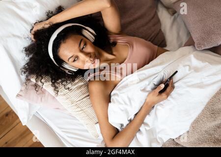Draufsicht auf eine lächelnde junge afrikanische Frau in Kopfhörern, die das Mobiltelefon im Bett hält Stockfoto