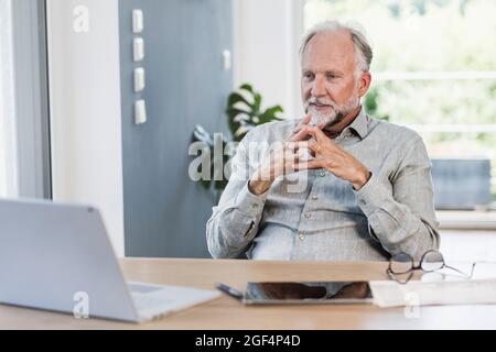 Geschäftsmann mit zusammengekrallten Händen und Blick auf den Laptop auf dem Schreibtisch im Heimbüro Stockfoto