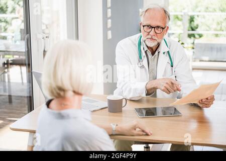 Männlicher Arzt, der einen medizinischen Bericht hält, während er eine Patientin am Schreibtisch ansieht Stockfoto