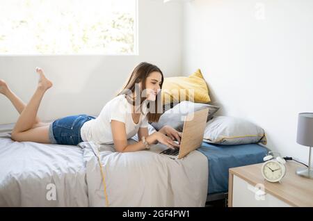 Junge Frau mit Laptop, während sie im Schlafzimmer zu Hause auf dem Bett liegt Stockfoto