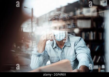 Reifer Mann, der eine schützende Gesichtsmaske trägt, liest Zeitung im Café, das durch das Glasfenster gesehen wird Stockfoto