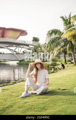Junge schöne blonde Frau trägt weiße Freizeitkleidung, Strohhut und Turnschuhe sitzen auf dem Gras im Park. Modisches, lässiges Sommeroutfit. Stockfoto