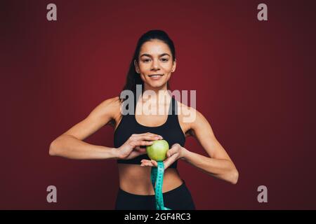 Lächelnde Athletin mit Apfel und Messband vor kastanienbraunem Hintergrund Stockfoto