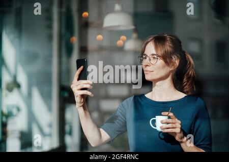 Junge Frau hält die Kaffeetasse, während sie auf das Mobiltelefon schaut Stockfoto