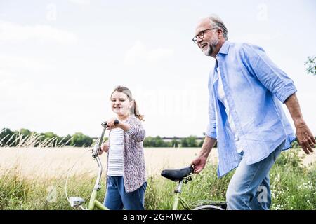 Lächelnder Großvater, der mit dem Fahrrad und der Enkelin auf der Wiese spazierengeht Stockfoto