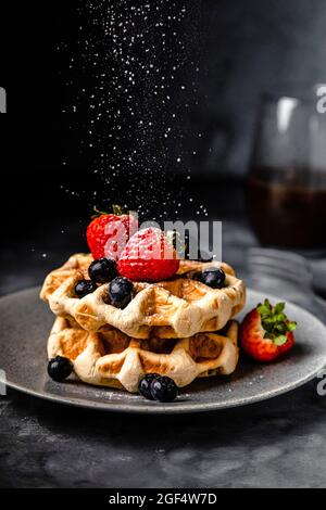 Puderzucker, der auf Teller mit frischen Waffeln mit Erdbeeren und Blaubeeren fällt Stockfoto