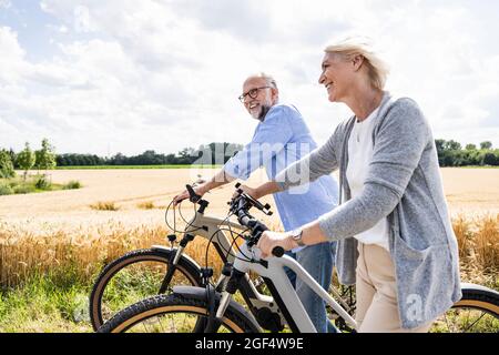 Lächelndes Paar mit Fahrrädern, das an sonnigen Tagen durch das Feld läuft Stockfoto