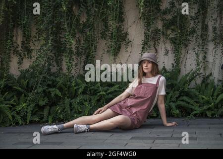 Teenager-Mädchen mit langen blonden Haaren auf der Straße sitzen. Modisches, lässiges Sommer- oder Frühlingsoutfit mit der Mütze. Hübsche junge Frau Stockfoto