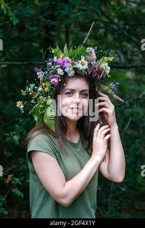 Portrait junge schöne Frau im Kranz von Wildblumen mit Farn im Wald. Blumenkräuterkranz auf dem Kopf der kaukasischen jungen Frau mit langem Braun Stockfoto