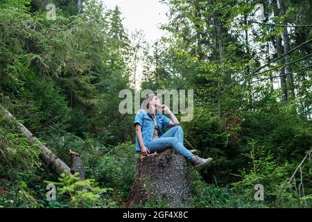 Frau, die im Wald im Park ruht. In Jeansanzug und Stiefeln gekleidet. Junge kaukasische Frau, die auf einem Baumstumpf sitzt und von Wald umgeben ist. Stockfoto