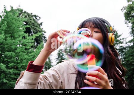 Junge Frau mit Kopfhörern, die im öffentlichen Park Blasen bläst Stockfoto