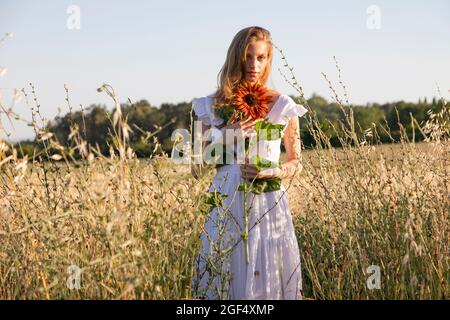 Junge blonde Frau hält Sonnenblume, während sie auf dem Feld steht Stockfoto