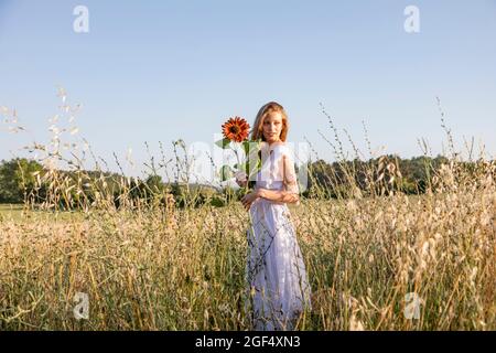 Junge Frau schaut weg, während sie Sonnenblume auf dem Feld hält Stockfoto