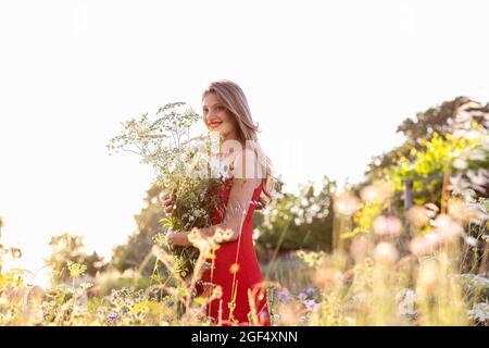 Lächelnde blonde Frau, die wegschaut, während sie den Blumenstrauß auf dem Feld hält Stockfoto