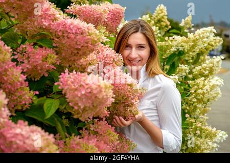 Glückliche attraktive Frau in den späten 40er Jahren, die ihren schönen Blumengarten genießt und zufrieden ihre blühenden Hortensien- und Hibiskusblüten in sich sieht Stockfoto