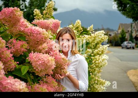 Glückliche attraktive Frau in den späten 40er Jahren, die ihren schönen Blumengarten genießt und zufrieden ihre blühenden Hortensien- und Hibiskusblüten in sich sieht Stockfoto