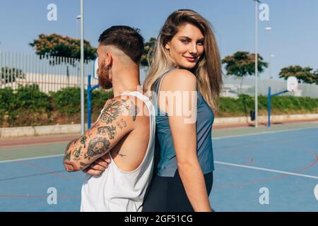 Lächelnde junge Frau, die am sonnigen Tag mit ihrem Freund Rücken an Rücken steht Stockfoto