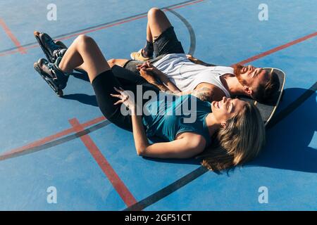Fröhliches junges Paar, das sich mit Skateboard ausruhte, während es auf dem Basketballplatz zusammenlag Stockfoto